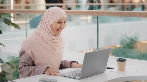Happy Young Arab Girl in Hijab Sitting at Table Answering Video Call on Laptop Joyfully Waving Hand