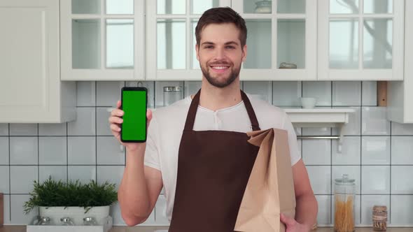 A Man Holds a Smartphone with a Green Screen in His Hand and Smiles While Standing in the Kitchen