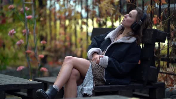 Thoughtful Young Woman Is Sitting on Bench at Autumn Day and Listening To Music By Headphones, Relax