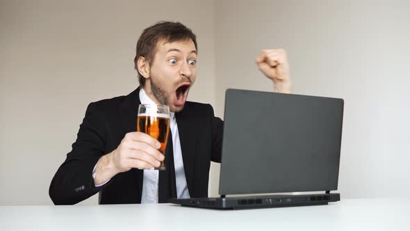 Happy businessman in suit sitting in front of laptop and holding glass of beer.