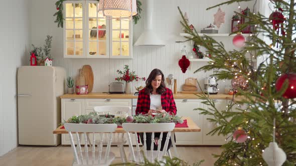 a Beautiful Woman in a Red Shirt Sits at a Decorated Christmas Table Against the Backdrop of a