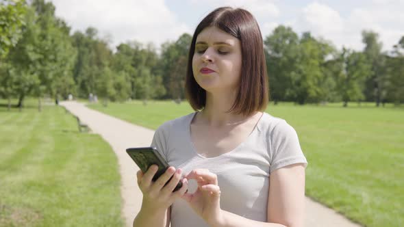 A Young Caucasian Woman Works on a Smartphone and Acts Frustrated on a Pathway in a Park