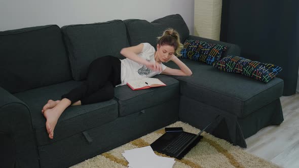Pretty woman makes notes in notebook while lying on couch at home in a modern apartment