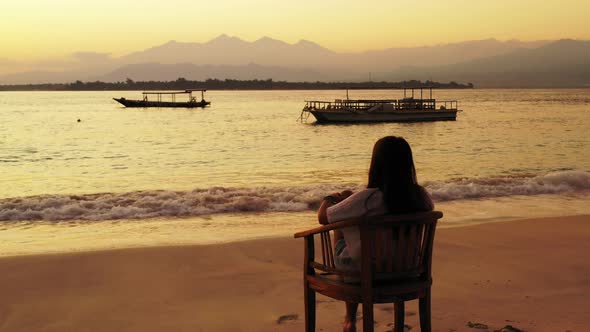 Melancholic girl sitting on chair over sandy beach, watching silhouette of boats floating on calm la