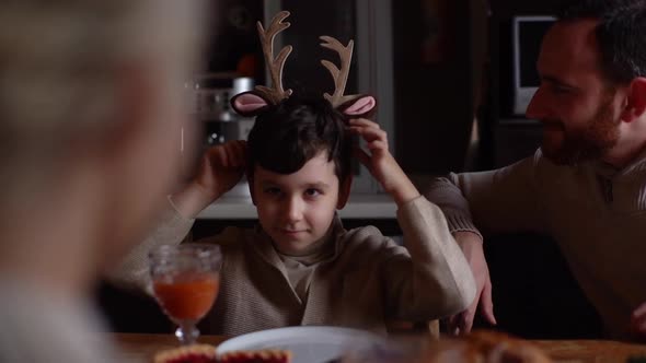 Joyful Little Child Boy Putting Toy Reindeer Antlers on Head Sitting at Festive Christmas Dinner