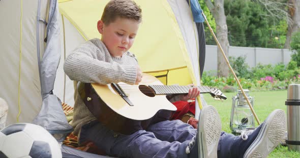 Caucasian boy sitting in tent in garden and playing guitar