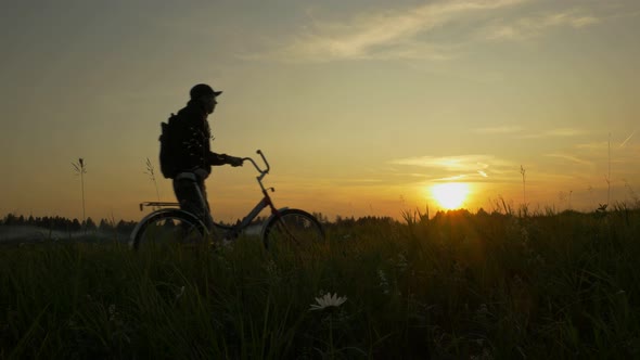 Male Traveler Walks Across the Field with a Bicycle on Sunset Background