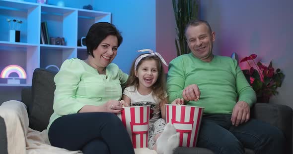 Funny Elder Family and Little Kid Granddaughter Sits on Sofa and Watching TV Eating Popcorn