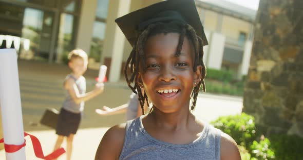 Video of happy african american boy wearing graduation hat and holding diploma in front of school