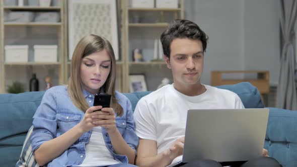 Young Couple Talking While Using Smartphone and Laptop at Home