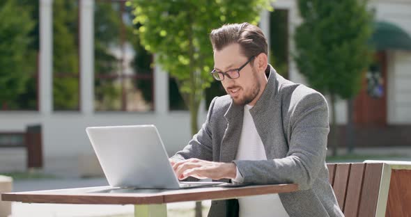 Young Man Wearing Glasses with Mustaches and a Beard is Sitting at the Table in the Square is Typing