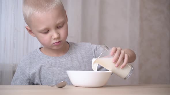 A light-haired little boy is pouring milk into a plate with corn flake.