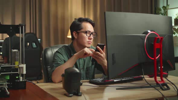 Asian Man Using Mobile Phone While Works On Personal Computer And 3D Printer In Home Office