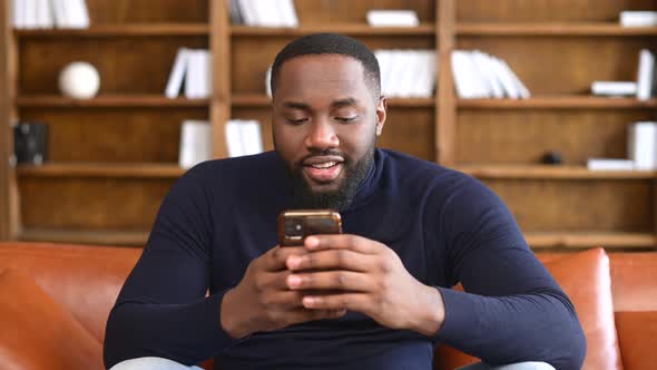 Excited Black Multiracial Man Sitting on the Brown Sofa Looking at the Phone