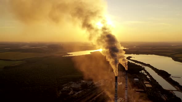 Industrial pipes with smoke in nature. Metallurgical plant among green fields at beautiful sunset.