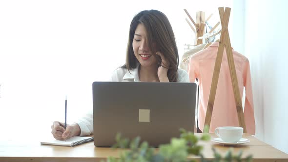 young smiling asian woman working on laptop while sitting in a living room at home using phone.