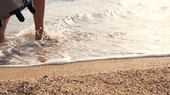 Close-up, Along Sandy Beach, in Foam of Surf, Female Legs Are Walking. Woman Carries a Travel