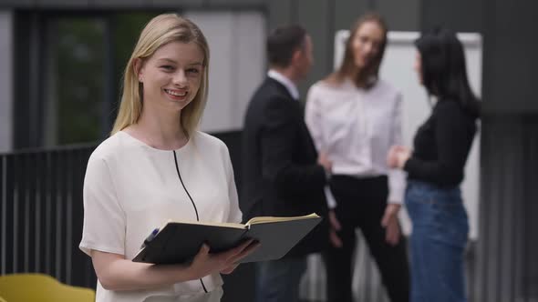 Medium Shot Portrait of Confident Charming Blond Woman Holding Paperwork Looking at Camera Smiling