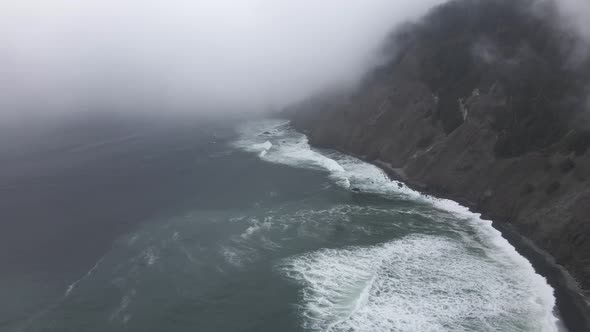 Fog hangs tight to steep cliffs as waves crash along a jagged coastline, aerial