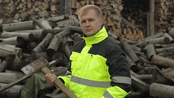 Lumberjack in Reflective Jacket. Man Woodcutter with Huge Axe. Sawn Logs, Firewood Background