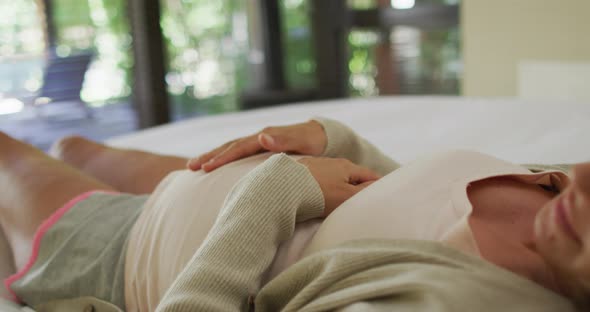 Midsection of caucasian pregnant woman touching belly and resting on bed