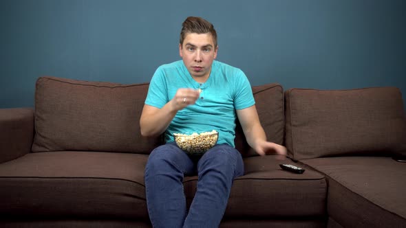 A Young Man Is Watching TV and Eating Popcorn. The Guy Is Watching TV Carefully. An Exciting Moment