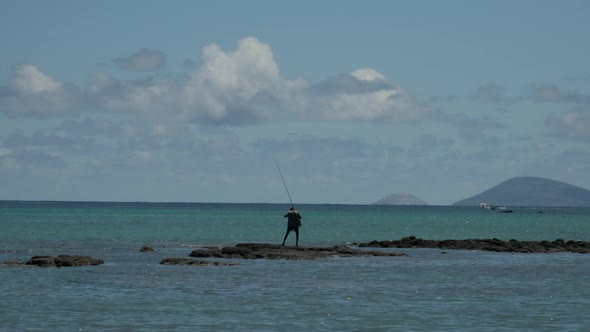 Swarthy mauritian man with large angle fishing on outstanding rock of pacific ocean. Wide shot.