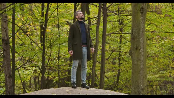 Smiling Caucasian Man with Gray Hair Standing Between Trees in the Autumn Forest and Looking