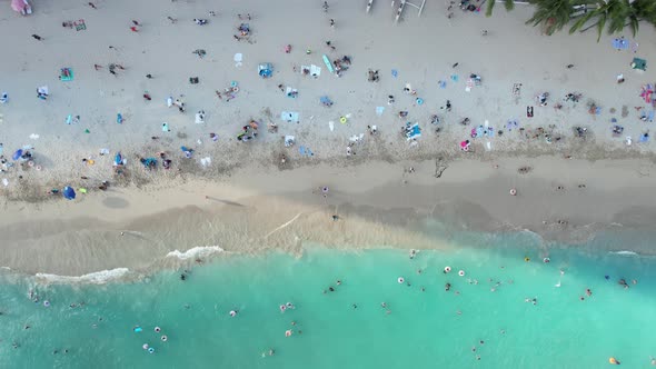 A drone hovers above a crowded tropical beach vacation destination as families enjoy sun, sand and s