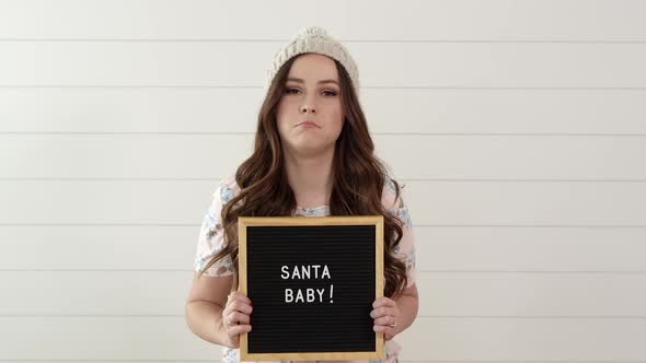 Woman frowning then smiling as she holds sign saying, Santa Baby