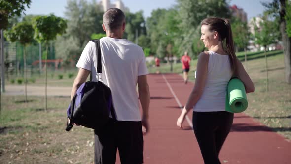 Back View of Positive Caucasian Man and Woman Walking on Sunny Sports Field and Talking. Camera