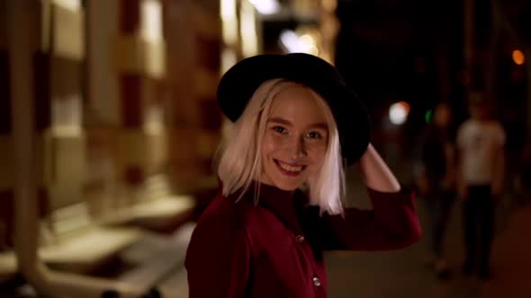 Hipster Blond Woman in Hat Turns Around with Beautiful Smile on Night City Street. Young Girl in