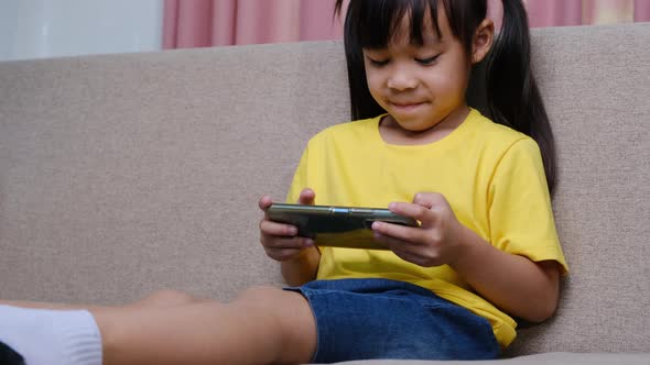 Cute little girl playing online games on her smartphone sitting on the sofa at home.