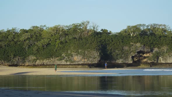 Two distant fisherman standing on sand bank during low tide with rock formation in background, Natad