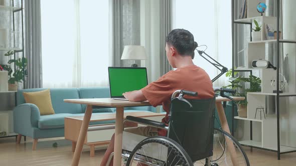 Back View Of Asian Boy Sitting In A Wheelchair While Using Laptop Computer With Green Screen At Home