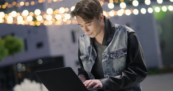 Young Man Uses a Laptop Sitting Outside in a Modern Residential Area, Evening in the City, Blurry