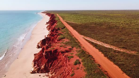 Drone tracking dirt road atop red ocean side cliffs with sandy beach and blue water