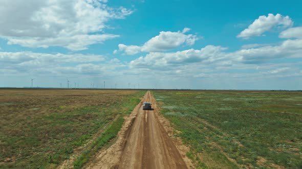 Drone Following Car Moving By Rustic Road Through Green Fields on Background of Wind Turbines