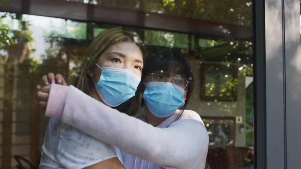 Asian mother and daughter looking through window with face masks embracing