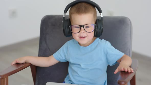 Cute Baby Boy Child in Glasses and Headphones Sitting Armchair Looking to Camera alt