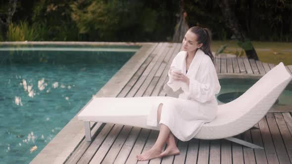 Woman in White Bathrobe Sits on Sunbed Near Pool, Drinks Coffee in Early Morning