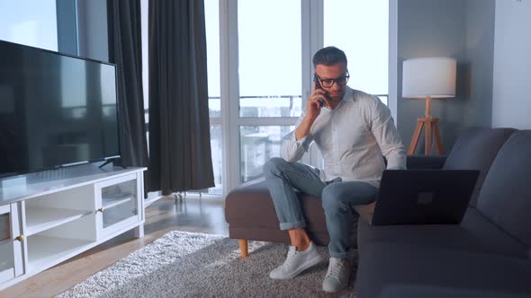 Man with Glasses Sitting on the Couch in a Cozy Room, Speaking on a Smartphone and Using Laptop