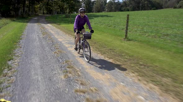 Mature woman in sunglasses and helmet biking on a gravel road lined with a wire fence on a sunny day