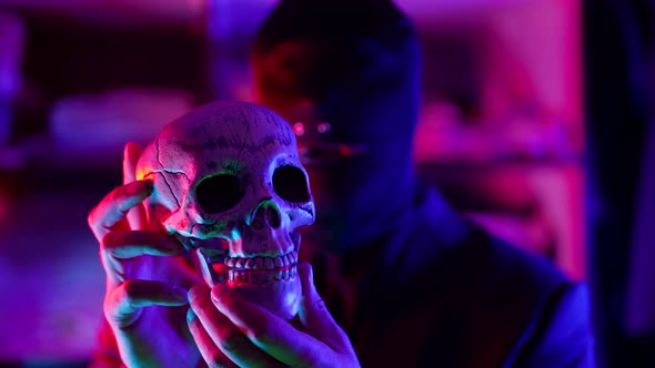 a Man Out of Focus and Wearing a Black Knitted Mask Without Eyes Holds a Skull in Front of Him and