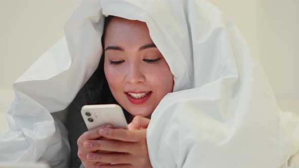 Cheerful Asian woman typing by phone under blanket