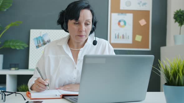 Slow Motion of Business Lady Wearing Headphones with Microphone Writing and Looking at Laptop Screen