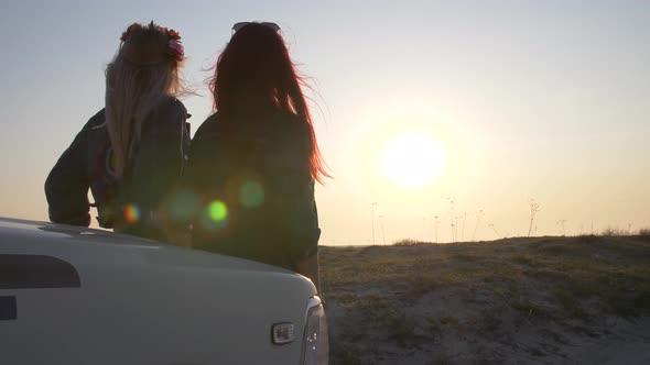 Couple of Young Women Is Sitting on the Bumper and Enjoy the Sunset