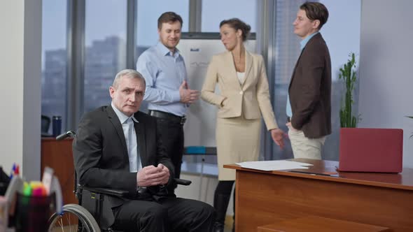 Depressed Caucasian Disabled Man in Wheelchair Looking at Camera As Blurred Coworkers Pointing and