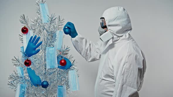 Doctior (Scientist) in Protective Suit, Goggle and Respirator Hangs a Ball on a Christmas Tree