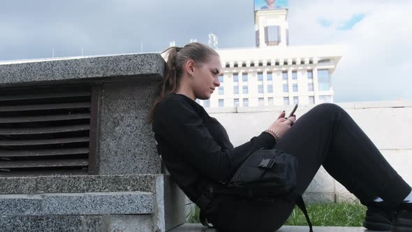 Young Woman Uses Smartphone While Sitting on the Parapet in the Street Park
